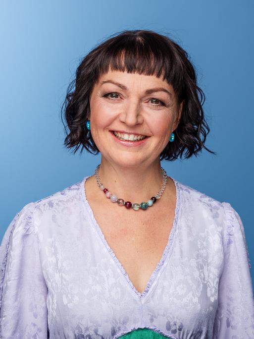 Doreen Bergmann lächelt in die Kamera. Sie trägt ein lavendelfarbenes Kleid und das dunkle Haar zu einem lockigen Bob geschnitten.