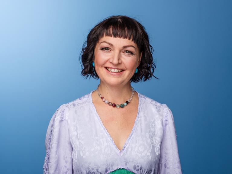 Doreen Bergmann lächelt in die Kamera. Sie trägt ein lavendelfarbenes Kleid und das dunkle Haar zu einem lockigen Bob geschnitten.