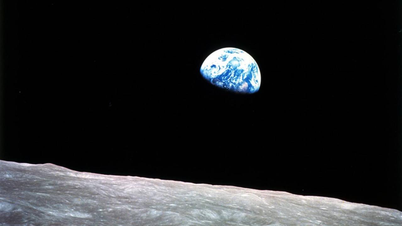 Beim Blick über eine steinige Ebene des Mondes erleuchtet die blau-weiß schimmernde Erde zur Hälfte im schwarzen Weltall.