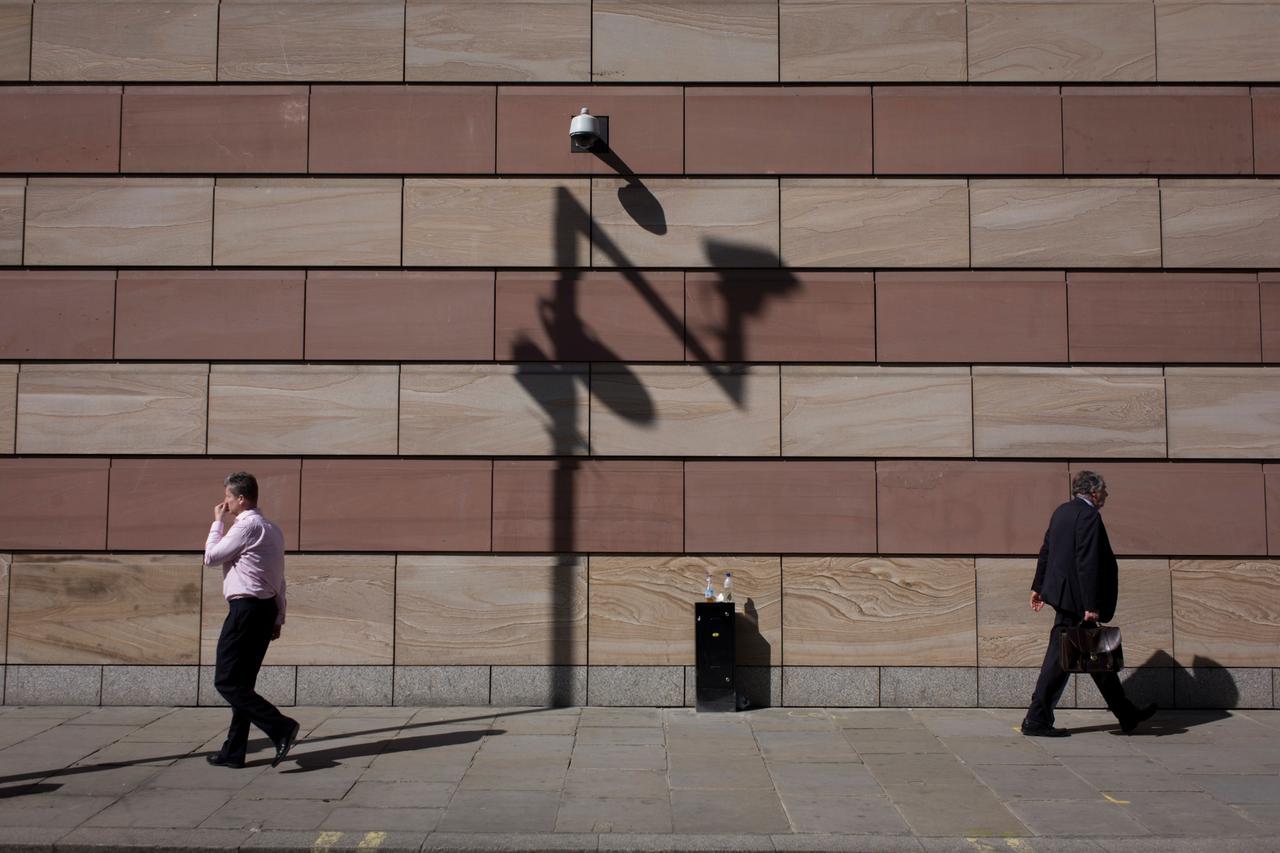Geschäftsleute passieren eine Überwachungskamera, die einen großen Schatten an die Wand eines modernen Bürogebäudes in der Finanzviertel City of London wirft.

