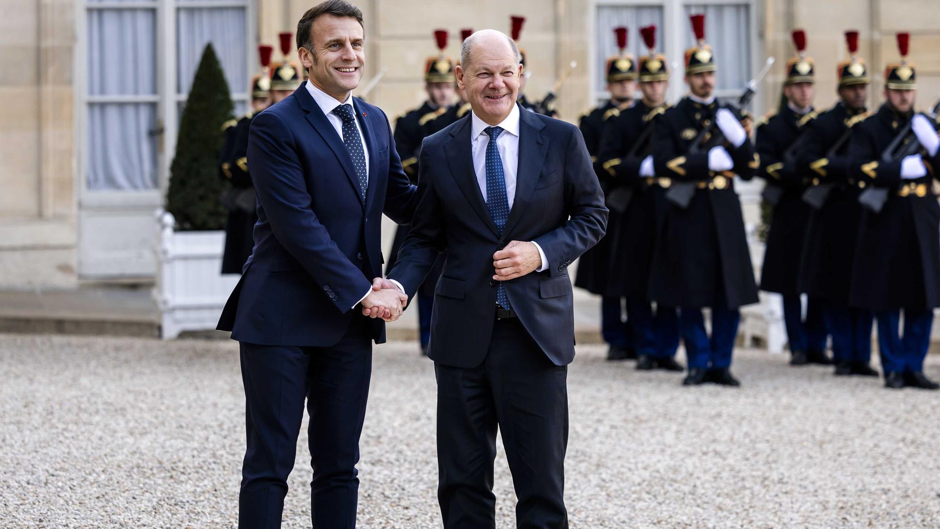 Bundeskanzler Olaf Scholz wird von Frankreichs Präsident Emmanuel Macron vor dem Elysee-Palast in Paris mit einem Handschlag begrüßt. Beide lächeln, im Hintergrund sind franzöische Soldaten zu sehen. 