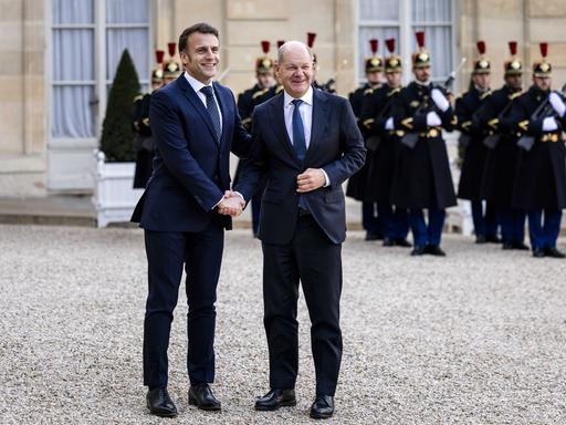 Bundeskanzler Olaf Scholz wird von Frankreichs Präsident Emmanuel Macron vor dem Elysee-Palast in Paris mit einem Handschlag begrüßt. Beide lächeln, im Hintergrund sind franzöische Soldaten zu sehen. 