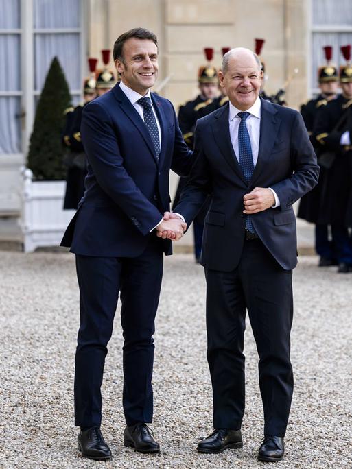 Bundeskanzler Olaf Scholz wird von Frankreichs Präsident Emmanuel Macron vor dem Elysee-Palast in Paris mit einem Handschlag begrüßt. Beide lächeln, im Hintergrund sind franzöische Soldaten zu sehen. 