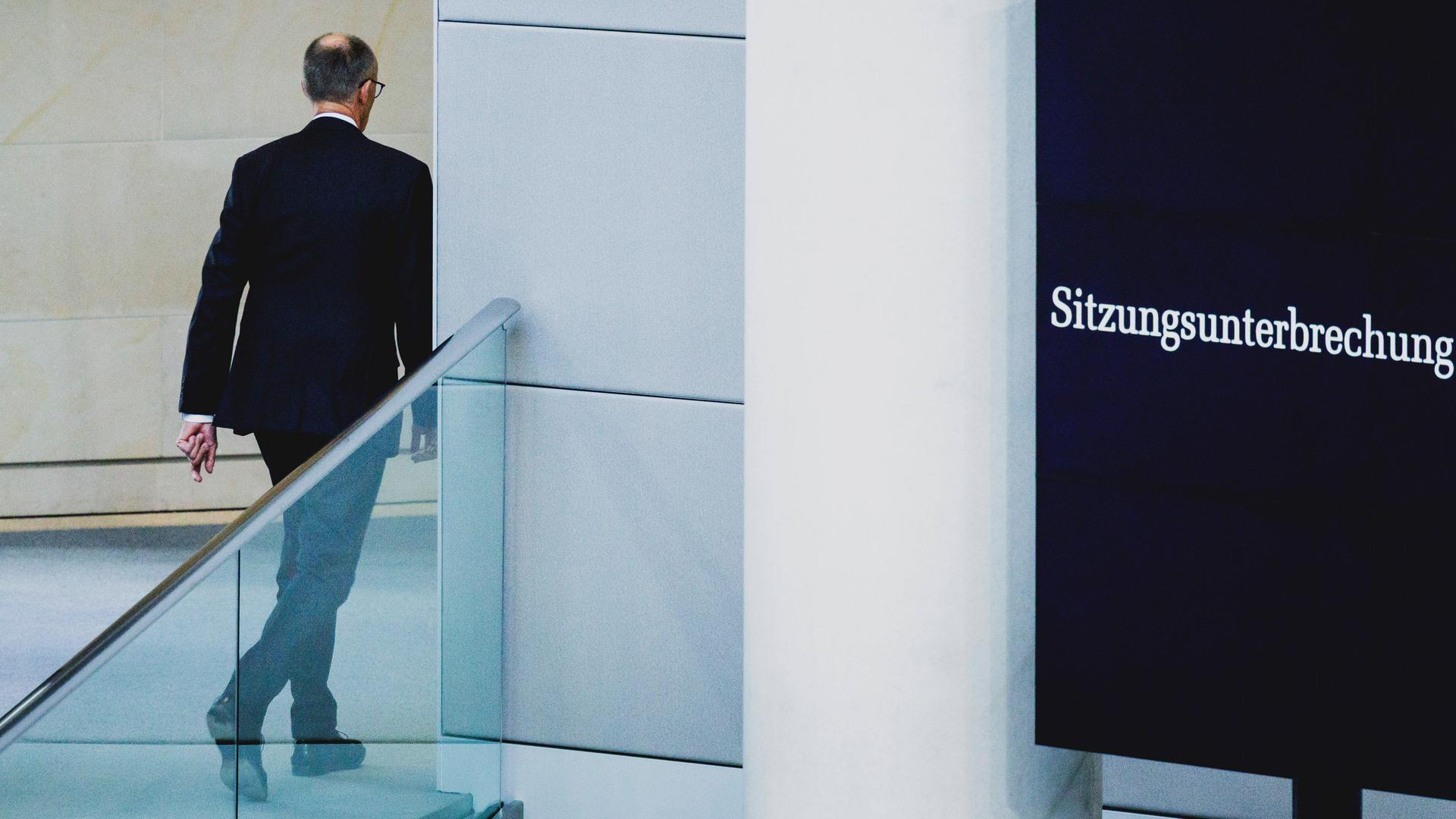 Friedrich Merz verlässt das Plenum des Bundestags, er ist von hinten zu sehen. Daneben zeigt ein Monitor "Sitzungsunterbrechung" an.