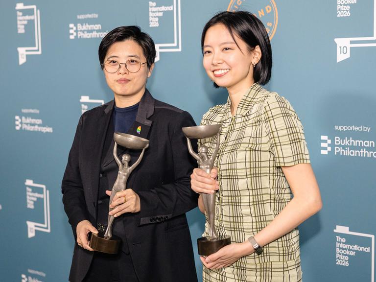 Zwei Frauen stehen nebeneinander und lächeln. Sie halten jeweils eine Statuette in silber.
