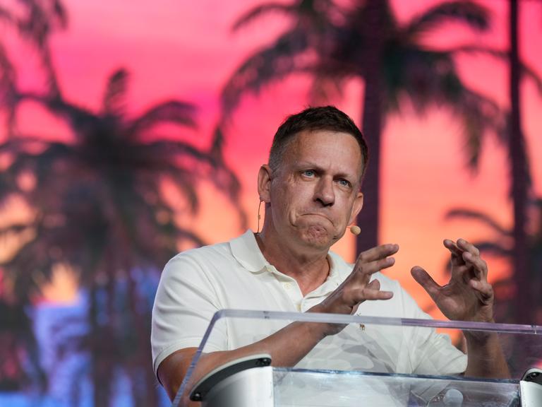 Peter Thiel steht an einem Redepult und gestikuliert mit den Händen. 