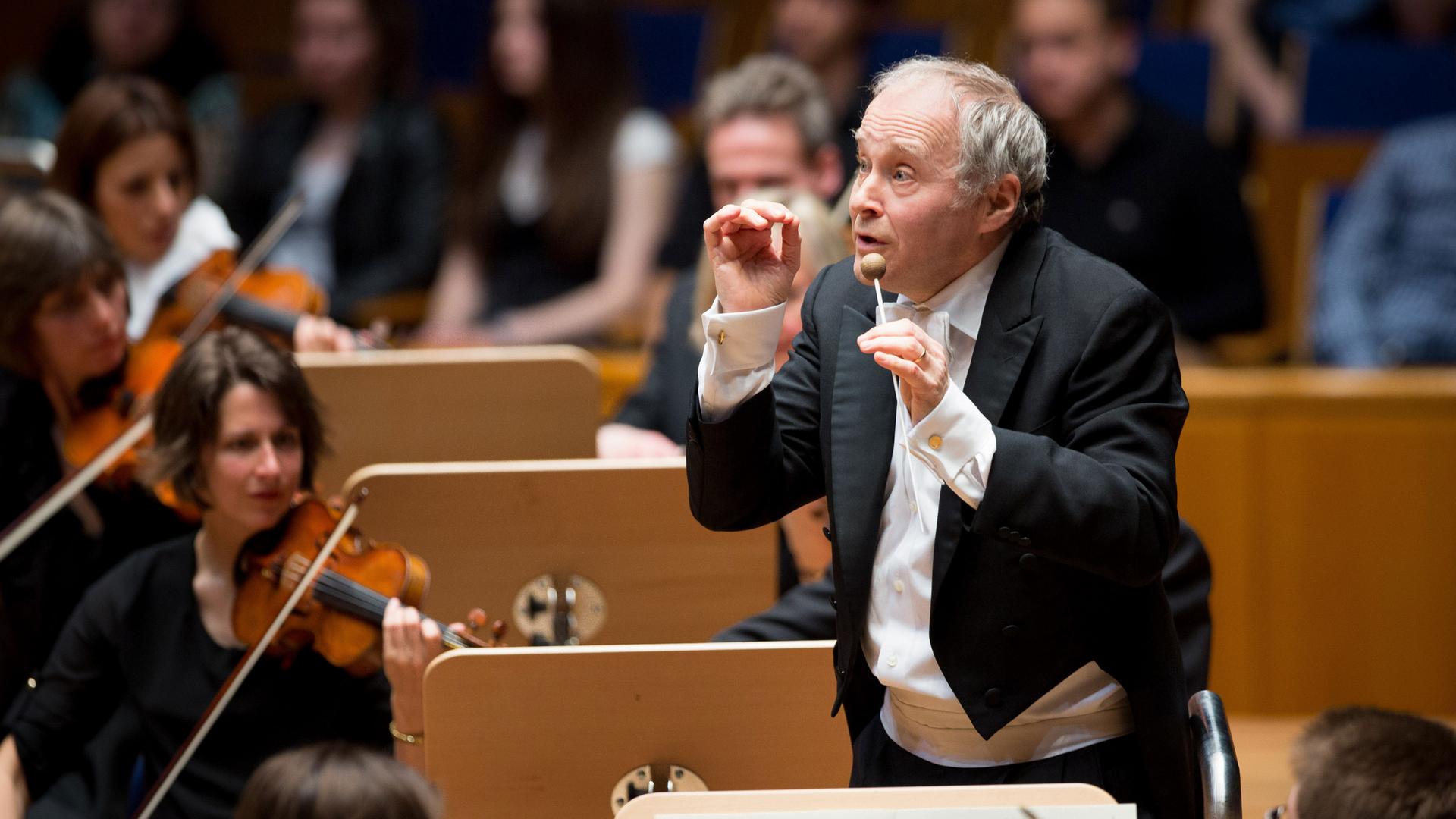 Der Dirigent Adam Fischer dirigiert vor einem Orchester, hinter ihm sind Notenpulte und mehrere Geigerinnen zu sehen. Der Dirigent Adam Fischer dirigiert vor einem Orchester, hinter ihm sind Notenpulte und mehrere Geigerinnen zu sehen.