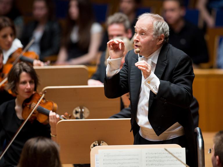 Der Dirigent Adam Fischer dirigiert vor einem Orchester, hinter ihm sind Notenpulte und mehrere Geigerinnen zu sehen.