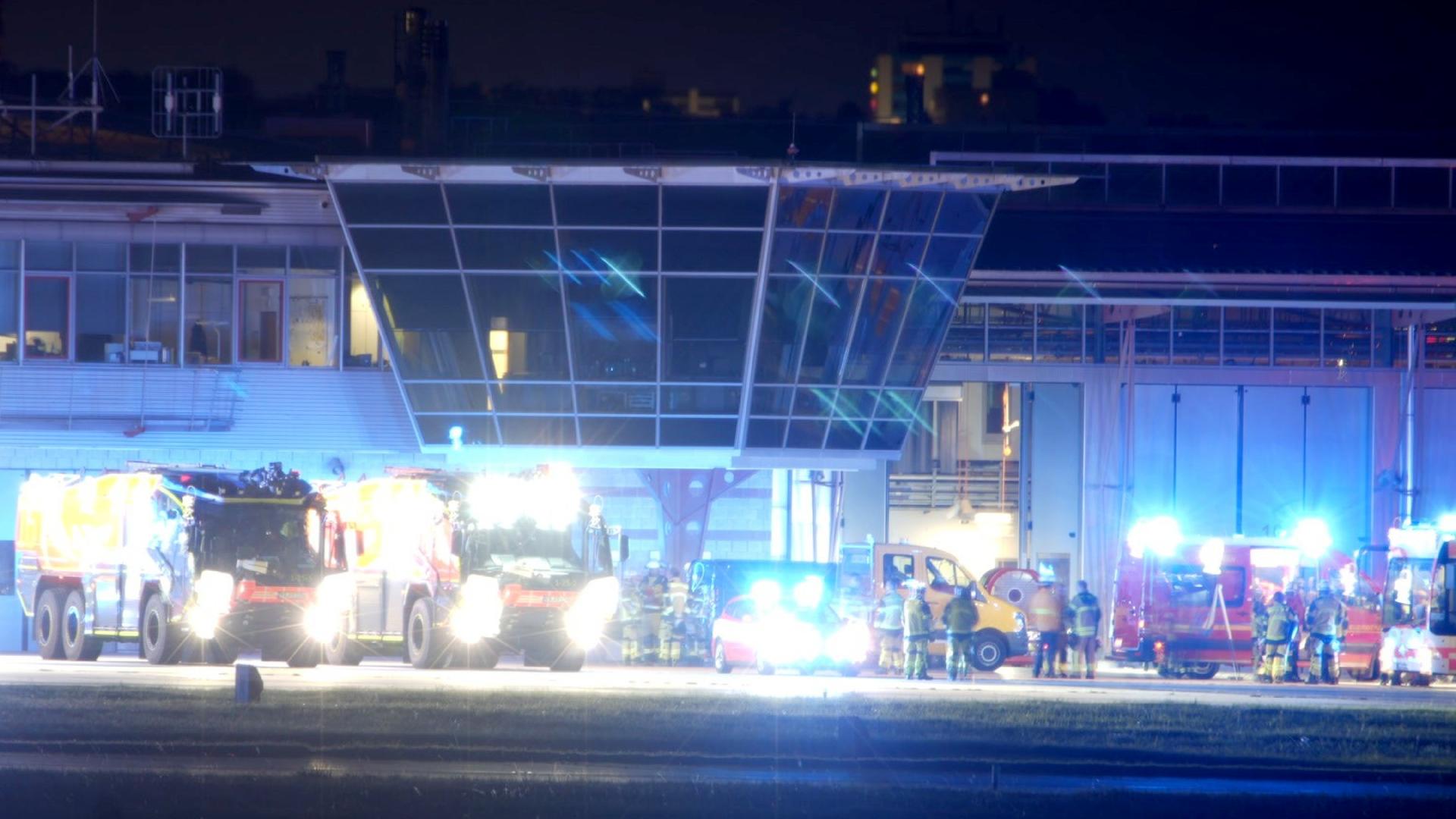 Vor einem Flughafen-Gebäude stehen Fahrzeuge von Feuerwehr und anderen Einsatzkräften mit hell leuchtenden Scheinwerfern und Blaulichtern.