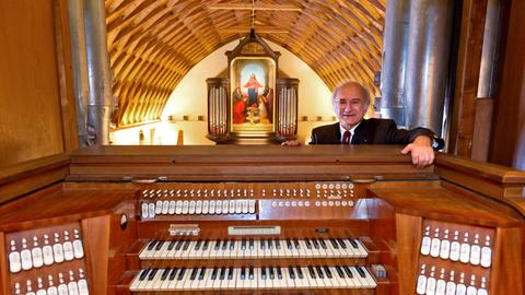Sixtus Lampl steht hinter einem Spieltisch einer großen Orgel. Hinter ihm eröffnet sich ein Raum mit hölzerner, gewölbten Decke mit Blick auf eine weitere Orgel, die mit einem großen Jesus-Bild geschmückt ist.