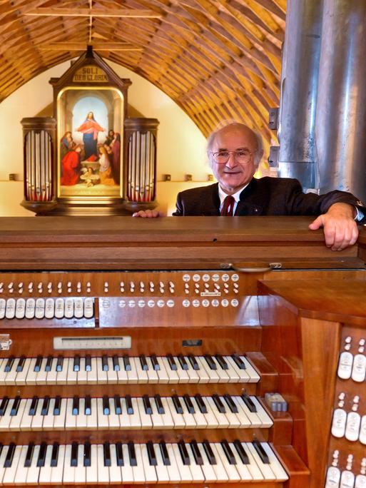 Sixtus Lampl steht hinter einem Spieltisch einer großen Orgel. Hinter ihm eröffnet sich ein Raum mit hölzerner, gewölbten Decke mit Blick auf eine weitere Orgel, die mit einem großen Jesus-Bild geschmückt ist.