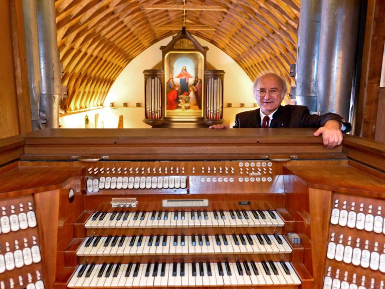 Sixtus Lampl steht hinter einem Spieltisch einer großen Orgel. Hinter ihm eröffnet sich ein Raum mit hölzerner, gewölbten Decke mit Blick auf eine weitere Orgel, die mit einem großen Jesus-Bild geschmückt ist.