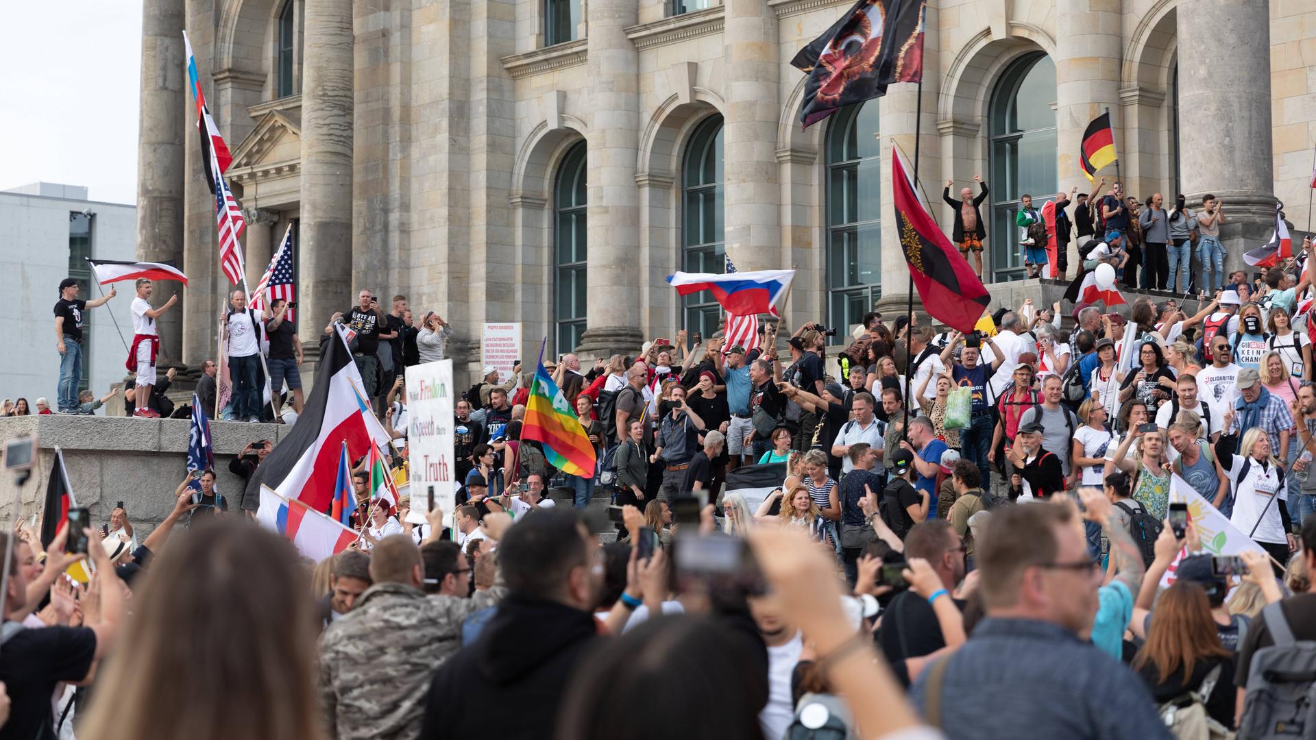 Blick auf den Reichstag, vor dem Hunderte Demonstrierende  gegen den Corona-Maasnahmen stehen. Dabei stürmen einige die Treppen des Reichstages. Eine Vielzahl von Flaggen ist zu sehen. Unter anderen die Regenbogenflagge, die Flagge der USA, die russische und die deutsche.
