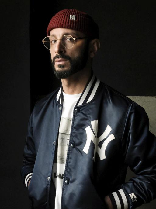 Behzad Karim Khani trägt eine Collegejacke und steht in einem dunklen Raum. Er blickt ernst in die Kamera.