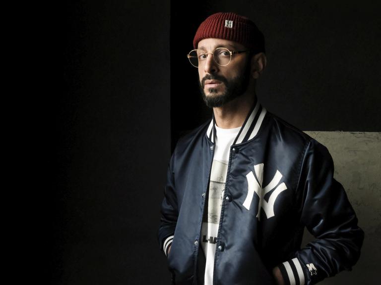 Behzad Karim Khani trägt eine Collegejacke und steht in einem dunklen Raum. Er blickt ernst in die Kamera.