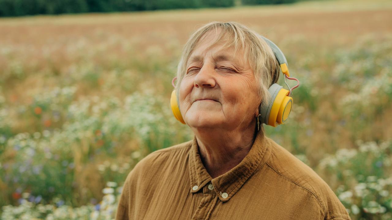 Eine ältere Frau steht inmitten eines Feldes, trägt große gelbe Kopfhörer und lächelt entspannt mit geschlossenen Augen. Sie wirkt versunken in die Musik.
