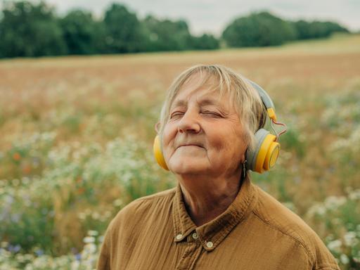 Eine ältere Frau steht inmitten eines Feldes, trägt große gelbe Kopfhörer und lächelt entspannt mit geschlossenen Augen. Sie wirkt versunken in die Musik.