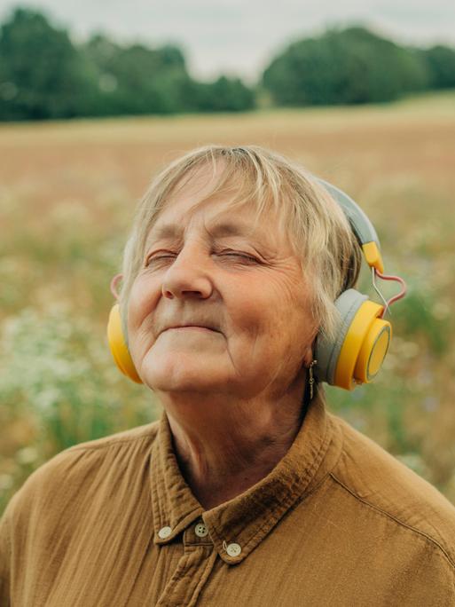 Eine ältere Frau steht inmitten eines Feldes, trägt große gelbe Kopfhörer und lächelt entspannt mit geschlossenen Augen. Sie wirkt versunken in die Musik.