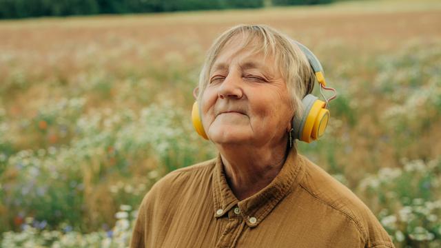 Eine ältere Frau steht inmitten eines Feldes, trägt große gelbe Kopfhörer und lächelt entspannt mit geschlossenen Augen. Sie wirkt versunken in die Musik.