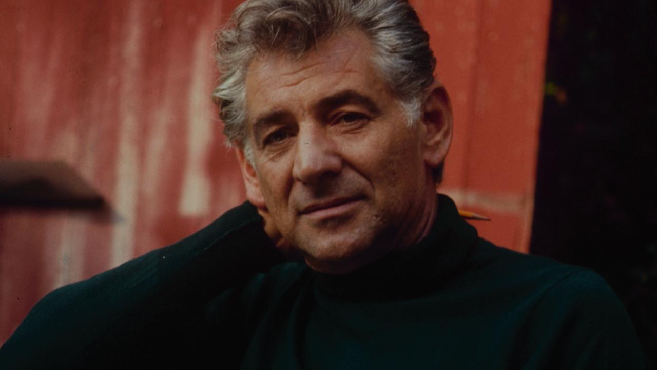 Leonard  Bernstein entspannt in einem Gartenstuhl und hält Blätter und seine Brille in der einen Hand, während er mit der anderen Hand seinen Kopf stützt. 