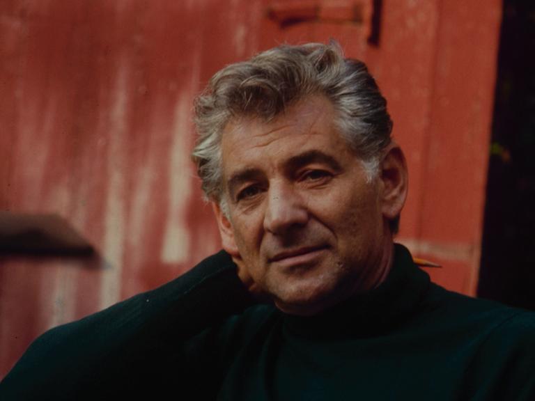 Leonard  Bernstein entspannt in einem Gartenstuhl und hält Blätter und seine Brille in der einen Hand, während er mit der anderen Hand seinen Kopf stützt. 