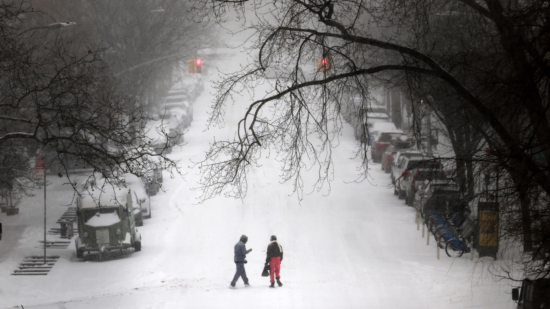 Starker Schneefall in New York: Zwei Personen gehen über eine verschneite Straße; am Rand stehen eingeschneite Autos.