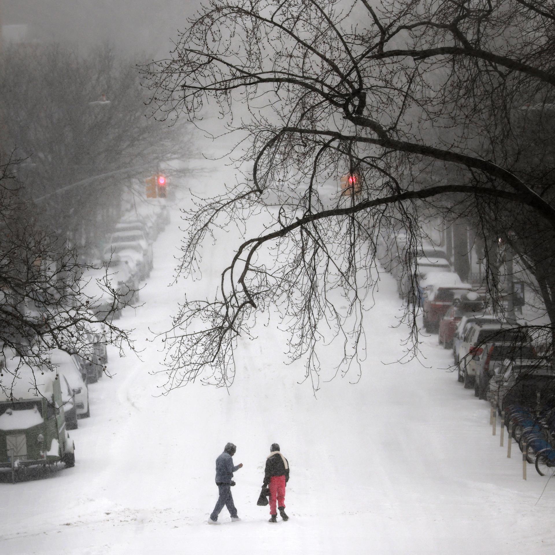 Starker Schneefall in New York: Zwei Personen gehen über eine verschneite Straße; am Rand stehen eingeschneite Autos.
