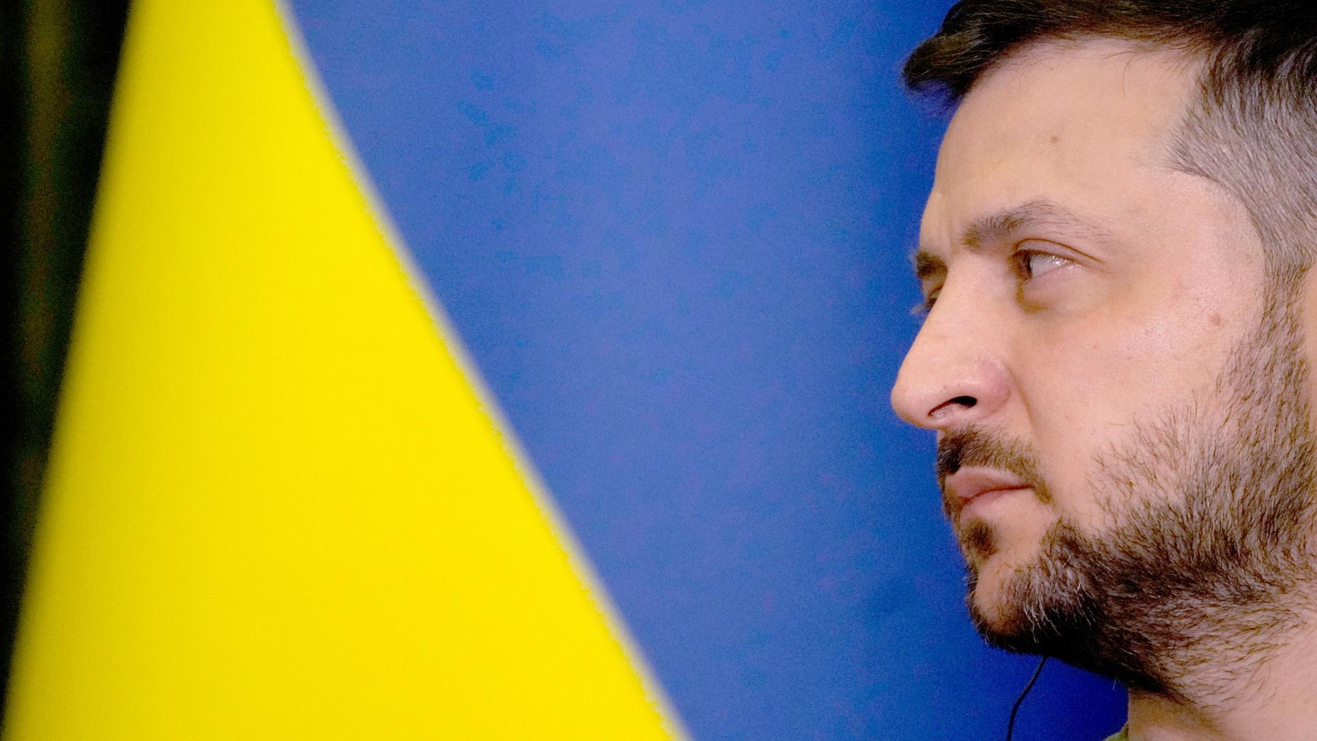 Der Präsident von der Ukraine Wolodymyr Selenskyj von der Seite. Dahinter die Flagge von der Ukraine in gelb und blau.
