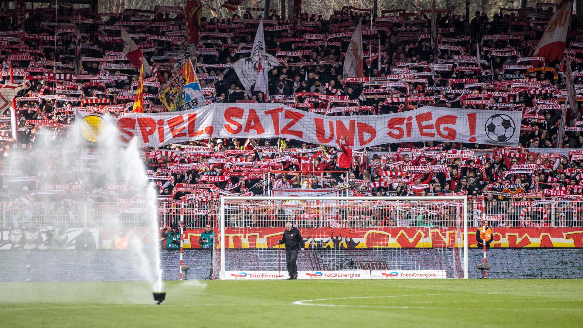 Fans von Union Berlin halten ein Banner mit der Aufschrift "Spiel, Satz und Sieg" in der Fankurve hoch.