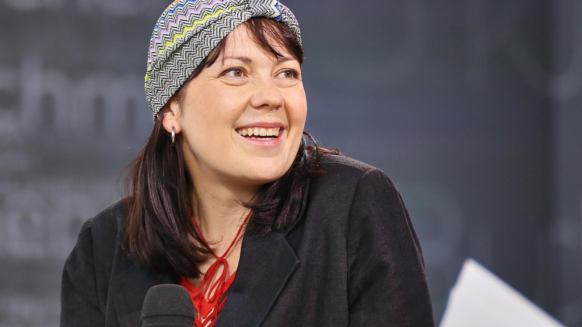 Dana von Suffrin sitzt mit einem Mikrofon auf einem Podium und lächelt ihr Gegenüber an. Sie trägt eine dekorative Kopfbedeckung.