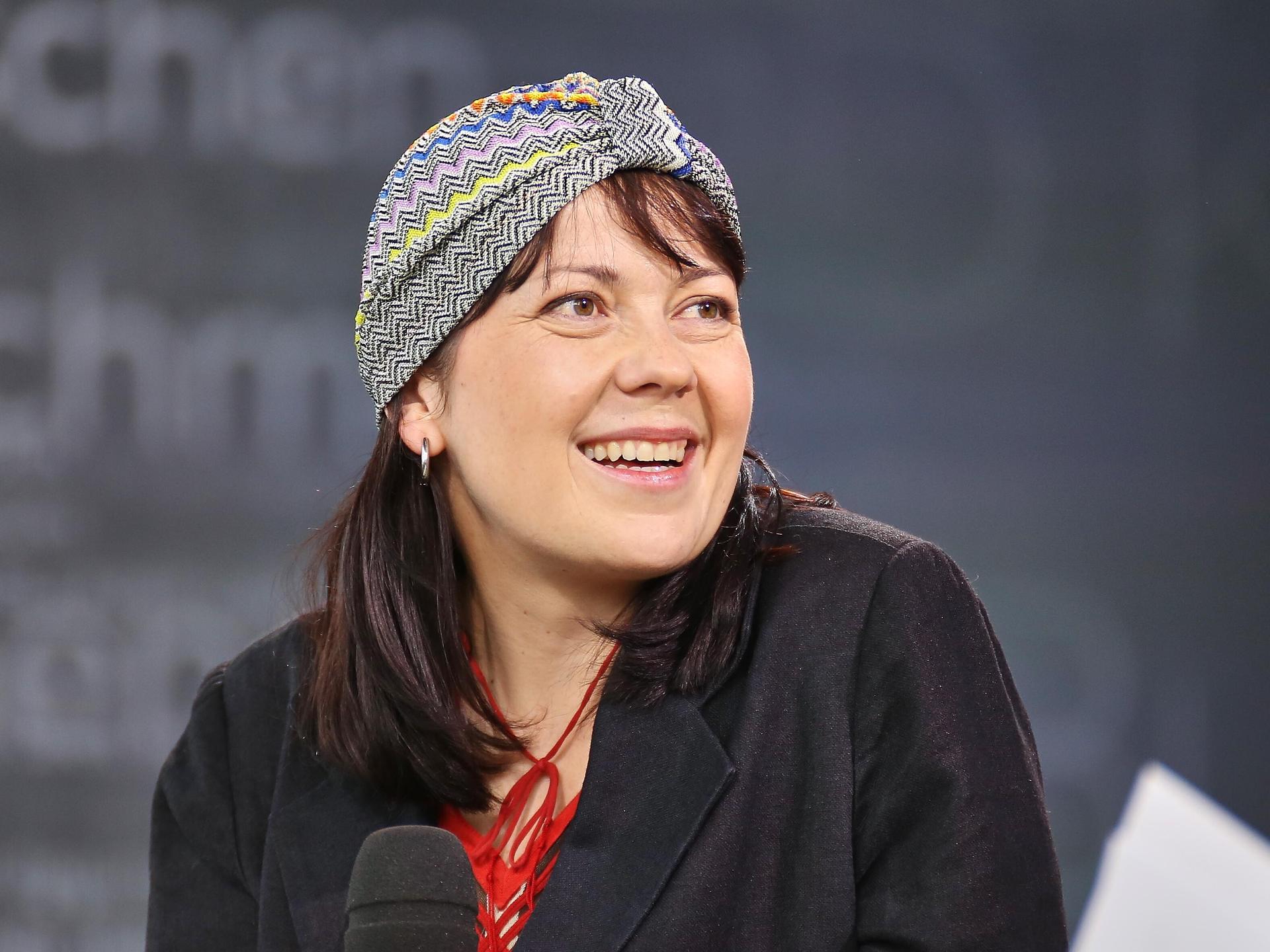 Dana von Suffrin sitzt mit einem Mikrofon auf einem Podium und lächelt ihr Gegenüber an. Sie trägt eine dekorative Kopfbedeckung.