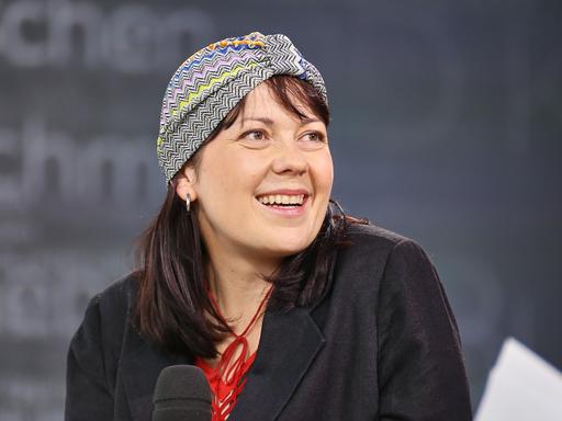 Dana von Suffrin sitzt mit einem Mikrofon auf einem Podium und lächelt ihr Gegenüber an. Sie trägt eine dekorative Kopfbedeckung.