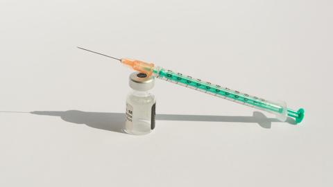 Nahaufnahme einer Impfstoffampulle und einer Injektionsspritze vor weißem Hintergrund. Ampulle und Spritze werfen einen harten Schatten. 