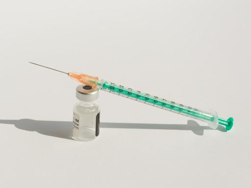 Nahaufnahme einer Impfstoffampulle und einer Injektionsspritze vor weißem Hintergrund. Ampulle und Spritze werfen einen harten Schatten. 