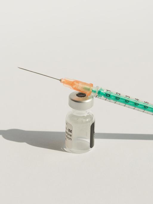 Nahaufnahme einer Impfstoffampulle und einer Injektionsspritze vor weißem Hintergrund. Ampulle und Spritze werfen einen harten Schatten. 