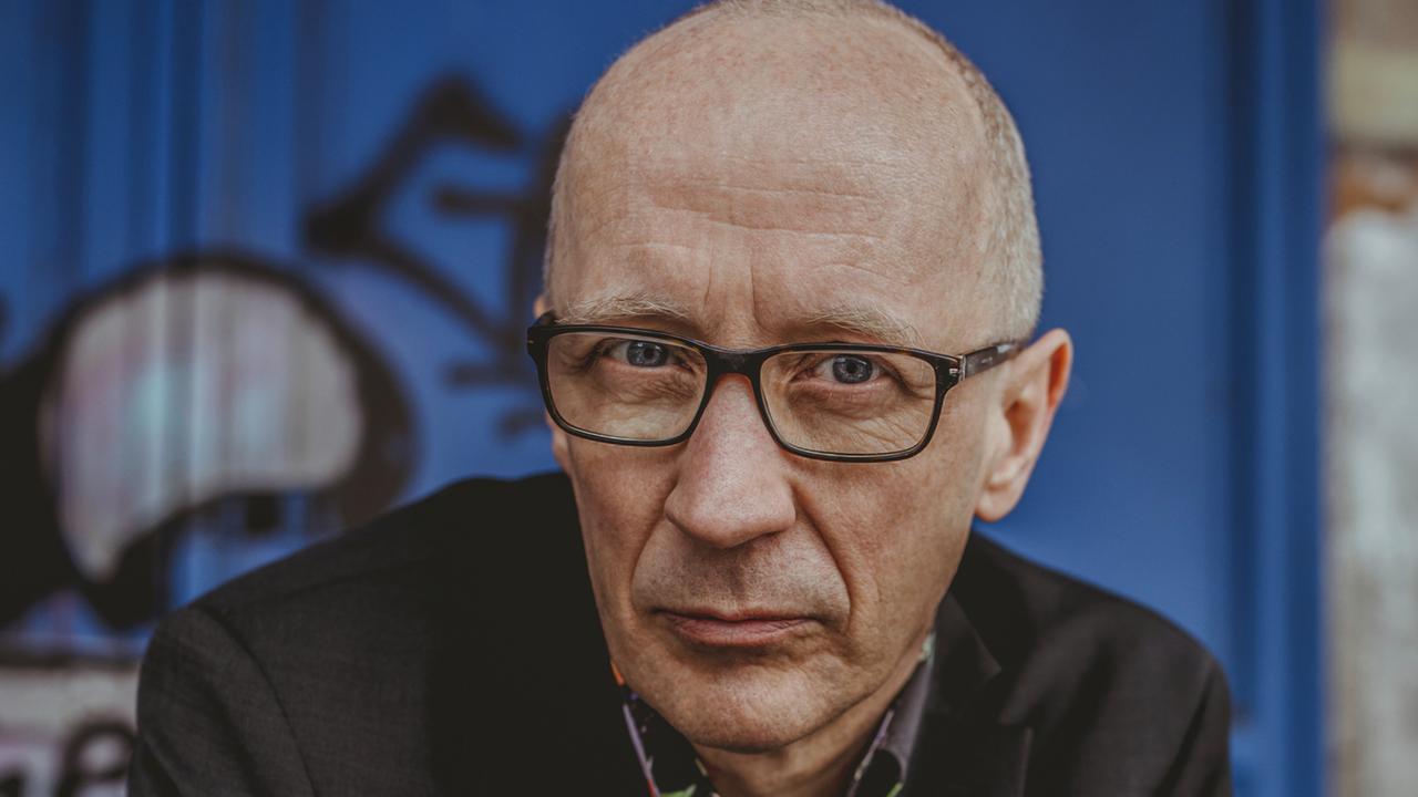 Ein Mann sitzt vor einer Wand mit Graffiti. Er trägt ein dunkles Sakko und ein buntes Hemd, er hat eine Glatze und trägt eine Brille. Ein Mann sitzt vor einer Wand mit Graffiti. Er trägt ein dunkles Sakko und ein buntes Hemd, er hat eine Glatze und trägt eine Brille.