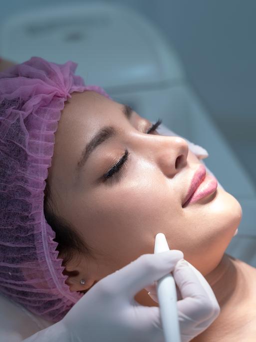 Eine Frau liegt für eine kosmetische Behandlung auf einer Liege. Sie trägt ein rosafarbenes Haarnetz.