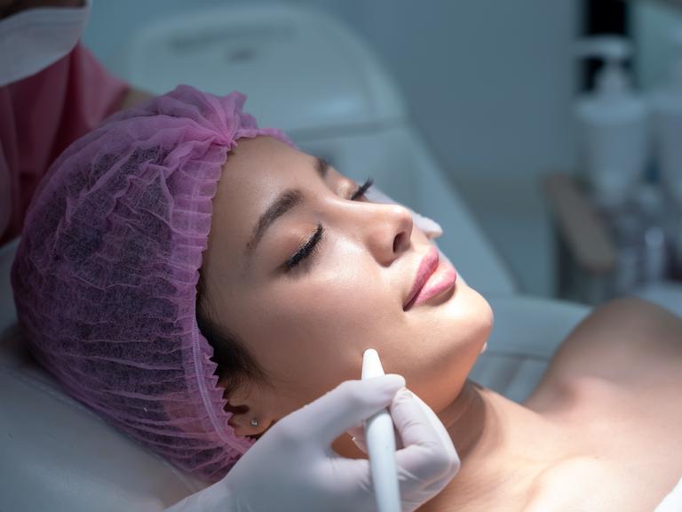 Eine Frau liegt für eine kosmetische Behandlung auf einer Liege. Sie trägt ein rosafarbenes Haarnetz.