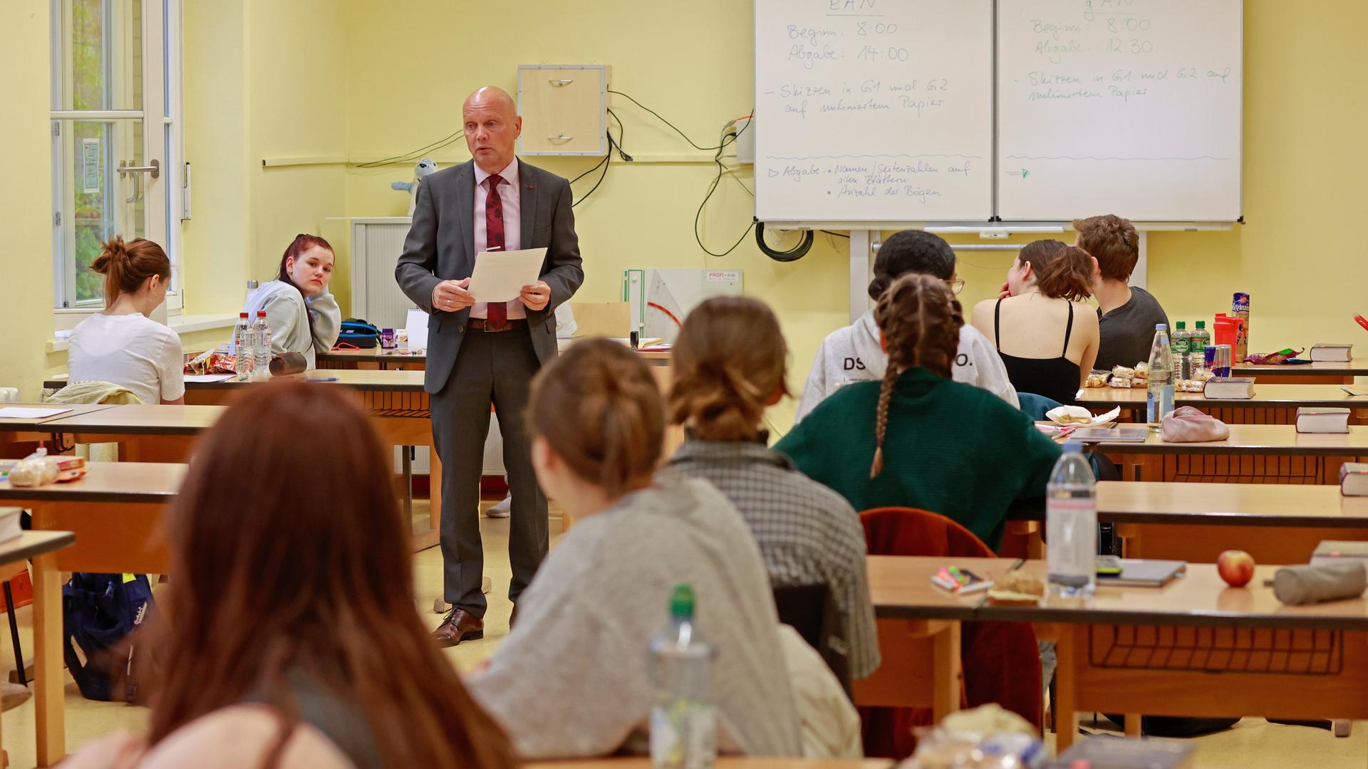 Schülerinnen und Schüler sitzen in eiinem Klassenraum an Pulten. In ihrer Mitte steht ein glatzköpfiger Mann mit Anzug und Krawatte.