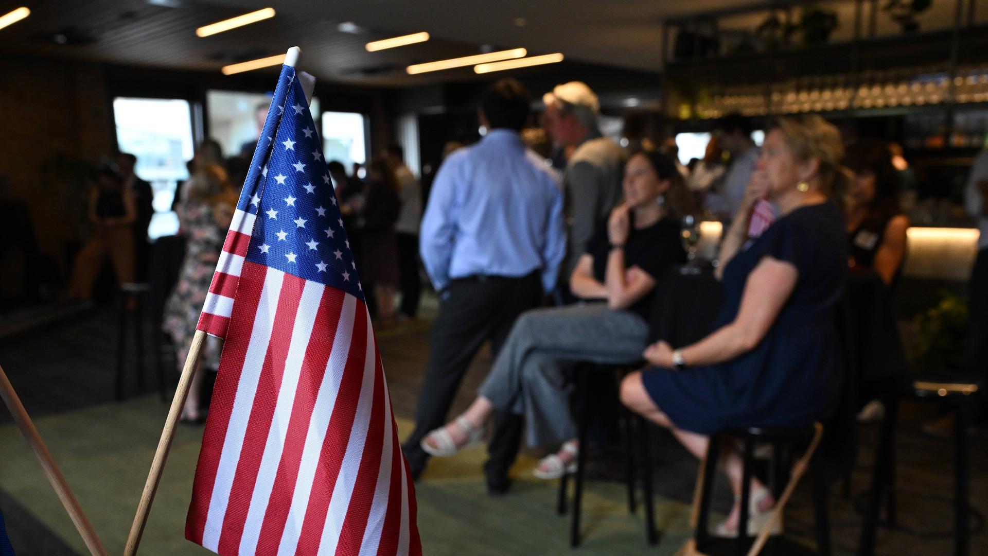 In den USA schauen in einer Bar Leute auf Bildschirme, auf denen die Wahlergebnisse eintrudeln. Im Vordergrund steht eine US-Flagge (6.11.2024)