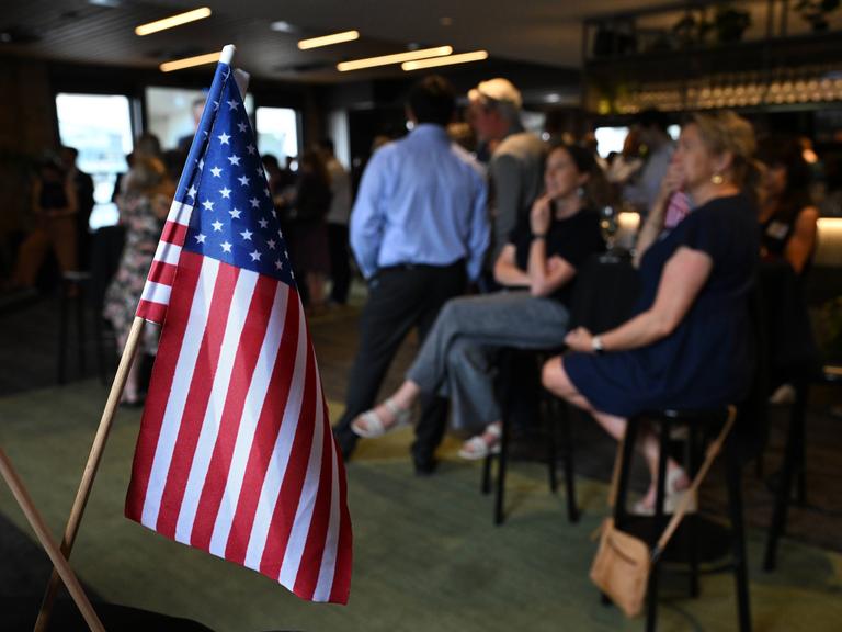 In den USA schauen in einer Bar Leute auf Bildschirme, auf denen die Wahlergebnisse eintrudeln. Im Vordergrund steht eine US-Flagge (6.11.2024)