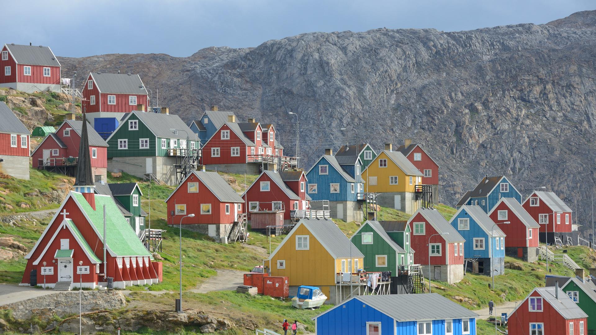 Der Ort Upernavik in Grönland. Bunte Häuser stehen in einer wunderschönen Naturlandschaft. Um die Häuser wächst grünes Grass. Im Hintergrund ein Fels.