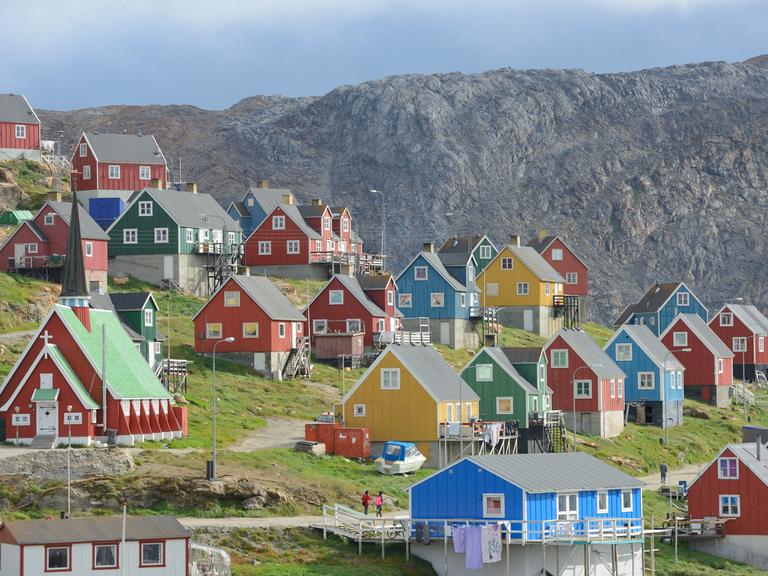 Der Ort Upernavik in Grönland. Bunte Häuser stehen in einer wunderschönen Naturlandschaft. Um die Häuser wächst grünes Grass. Im Hintergrund ein Fels.