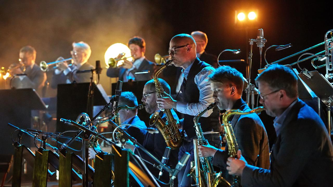 Ein Bläser-Ensemble mit Saxofonisten und Trompetern. Alle spielen auf ihren Instrumenten und stehen auf einer Bühne.