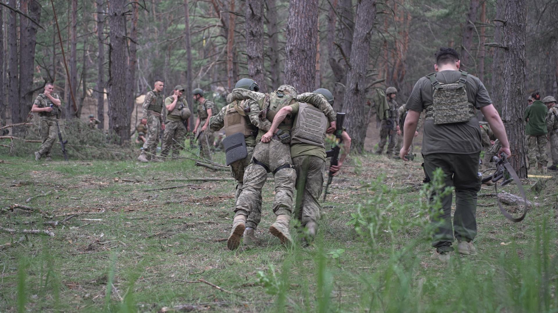 Ukrainische Soldaten trainieren in einem Waldstück einen Rettungseinsatz. Ein Soldat wird von seinen Kameraden zwischen den Schultern weggetragen.