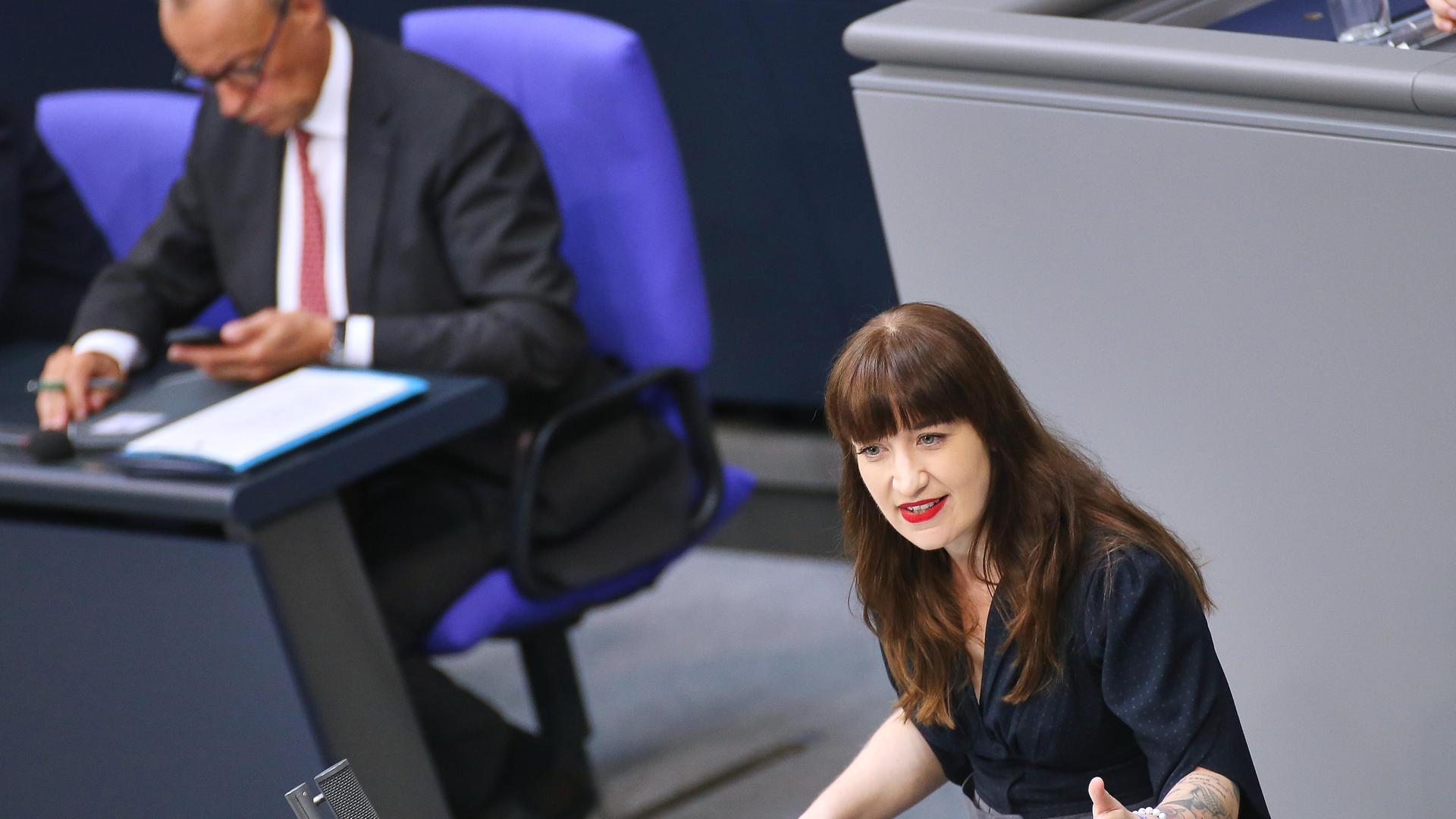 Linken-Chefin Heidi Reichinnek steht im Bundestag am Redepult und spricht. Im Hintergrund sitzt Kanzler Friedlich Merz im Plenum und blickt auf sein Handy. 
