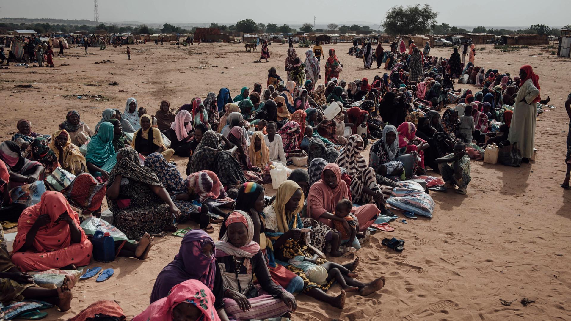 Eine große Gruppe Flüchtlinge sitzt in mehreren Reihen nebeneinander auf einer sandigen Fläche.