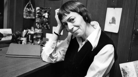 Schwarzweiß-Fotografie von Ursula K. Le Guin an ihrem Schreibtisch. Sie hat den Kopf auf ihre Hand gelehnt und lächelt freundlich in die Kamera