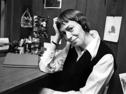 Schwarzweiß-Fotografie von Ursula K. Le Guin an ihrem Schreibtisch. Sie hat den Kopf auf ihre Hand gelehnt und lächelt freundlich in die Kamera