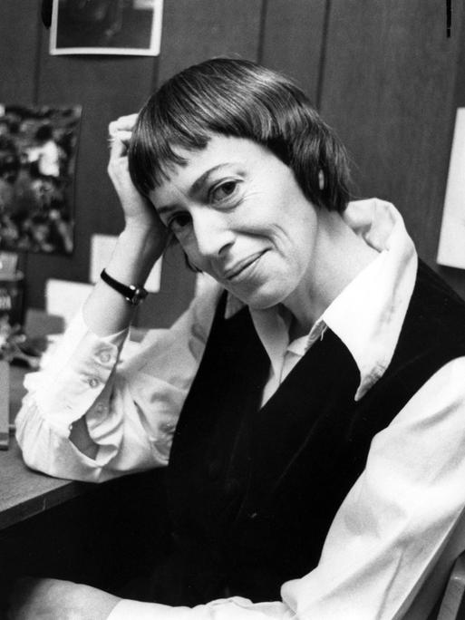 Schwarzweiß-Fotografie von Ursula K. Le Guin an ihrem Schreibtisch. Sie hat den Kopf auf ihre Hand gelehnt und lächelt freundlich in die Kamera